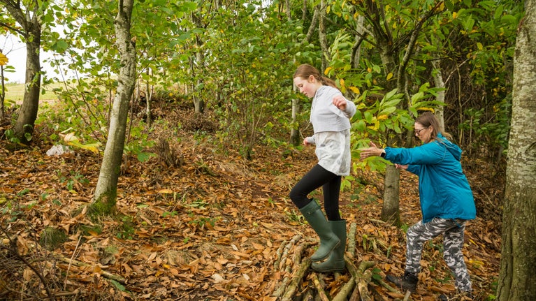 Woman and girl clambering uphill through woodland over logs and leaves